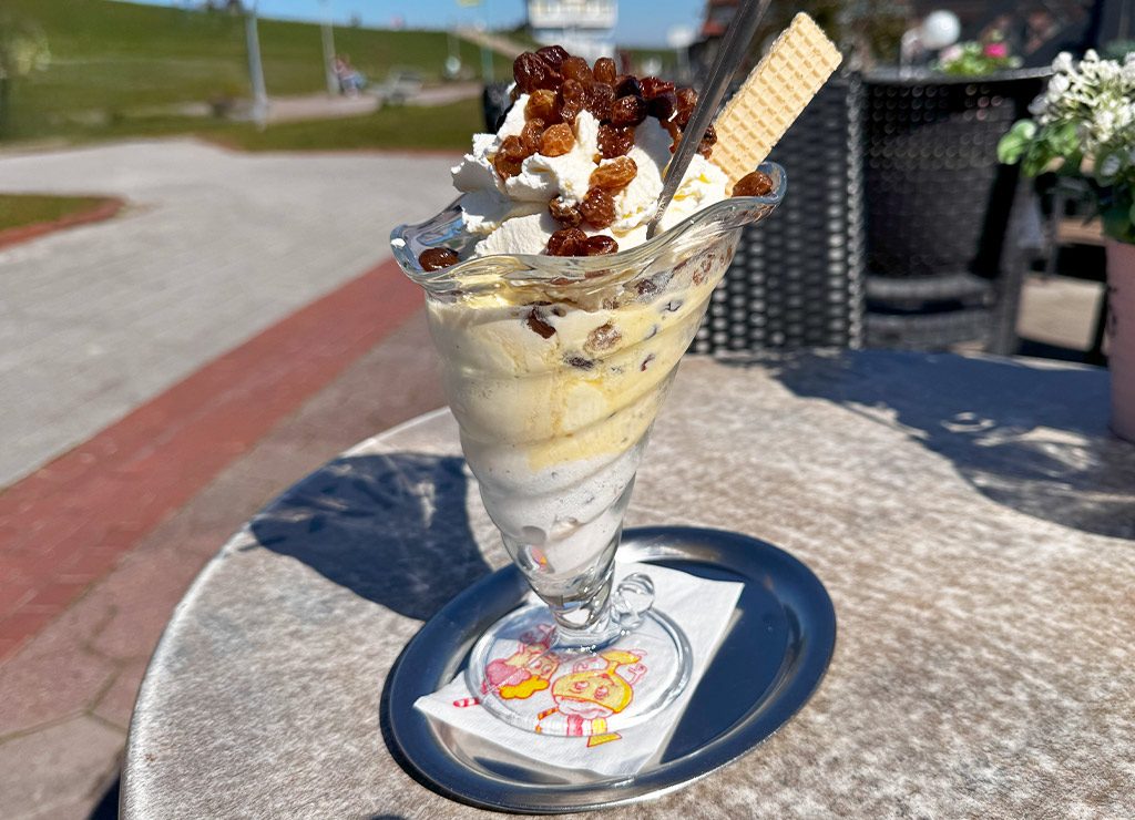 National Ice Cream Day – Tag des Eiscreme in den USA. Kuriose Feiertage – dritter Sonntag im Juli (c) 2025 Dietmar Giese – Bild 1 Großer Eisbecher mit Rosinen, Sahne und Waffel auf einem Tisch im Sonnenschein serviert.