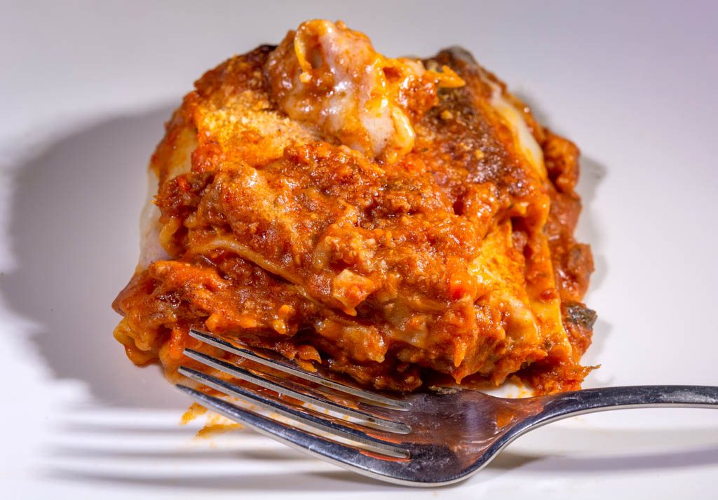 National Lasagna Day – Tag der Lasagne in den USA. Kuriose Feiertage - 29. Juli © 2023 Sven Giese – Bild 1 Ein Stück Lasagne mit viel Tomatensauce und Gabel auf weißem Teller angerichtet.