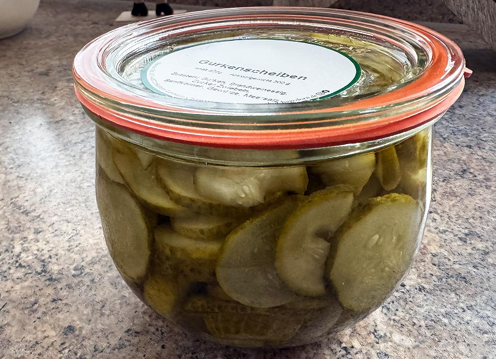 Ein Glas mit eingelegten Gewürzgurkenscheiben steht auf einer steinernen Küchenplatte im Tageslicht. 