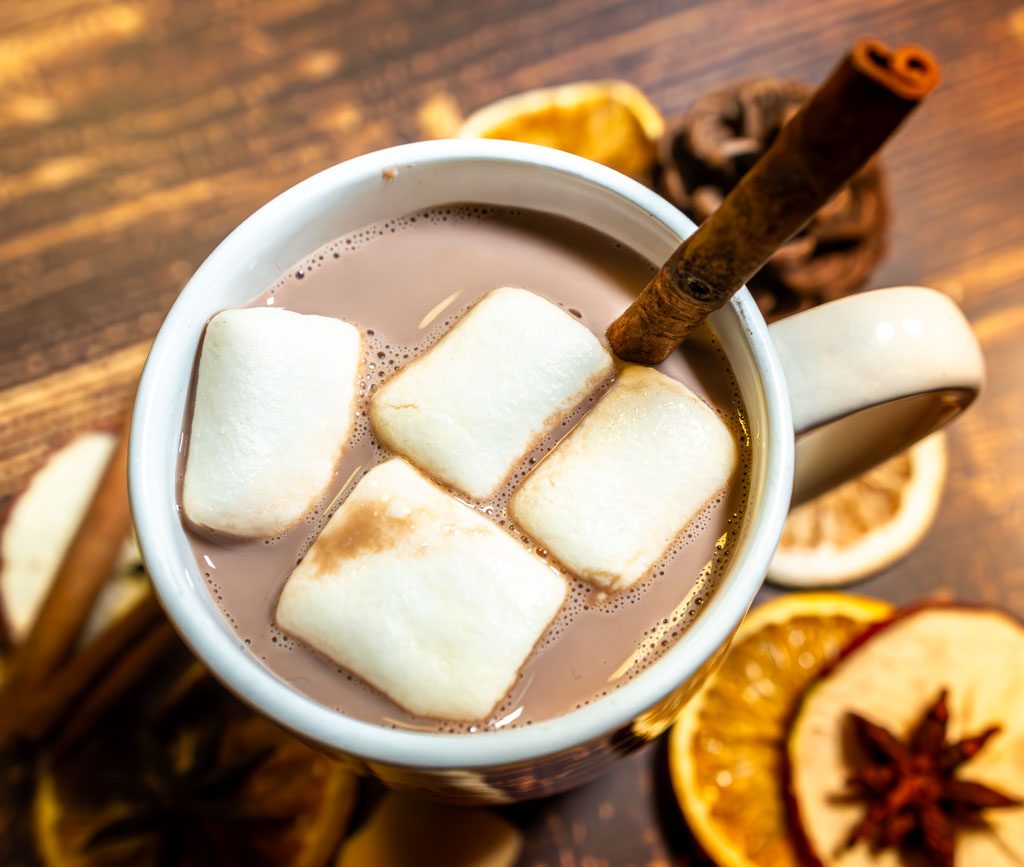 Weiße Tasse mit heißem Kakao, Marshmallows und Zimtstange, umgeben von getrockneten Orangenscheiben.