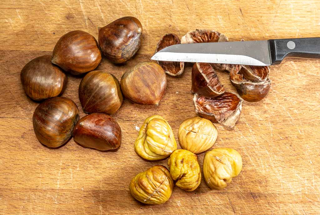 Frische und geschälte Maronen auf einem Holzbrett, daneben ein Messer und Schalenreste.