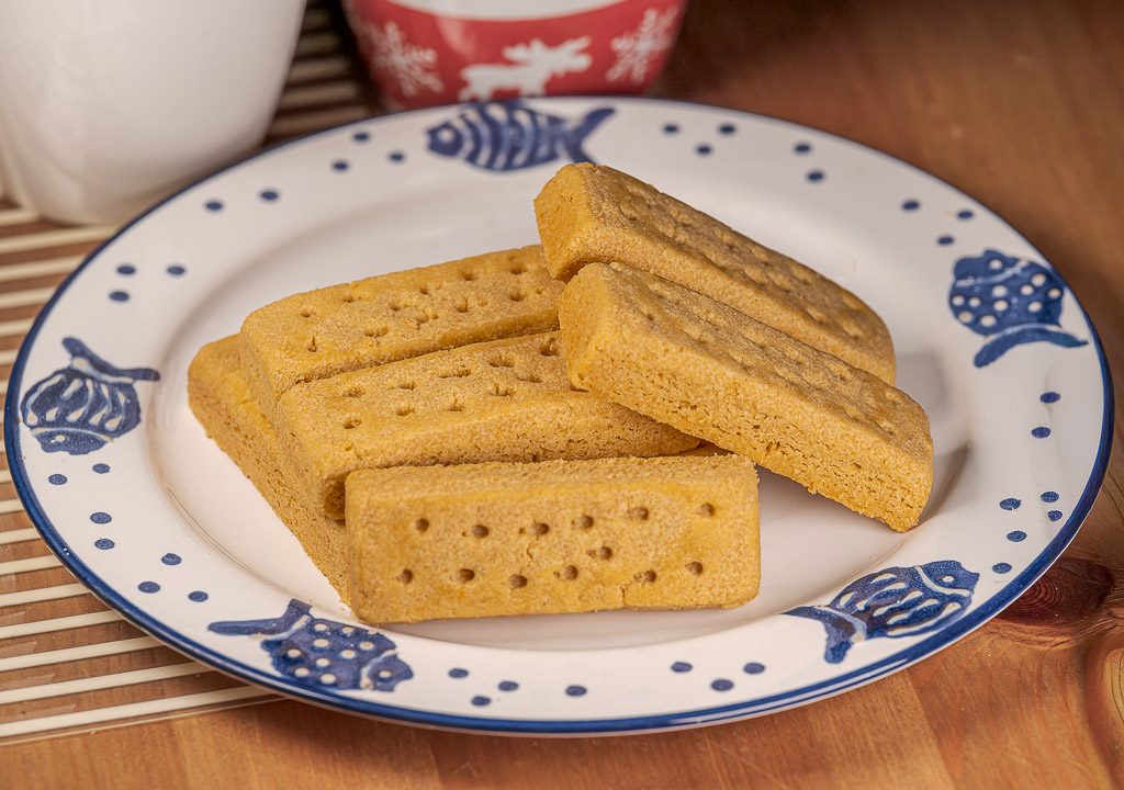 National Shortbread Day - Tag des schottischen Shortbread in den USA. Kuriose Feiertage - 6. Januar © 2026 Sven Giese - Bild 2 Mehrere Shortbread-Riegel auf gemustertem Teller, serviert neben Tasse auf Holztisch im warmen Licht.