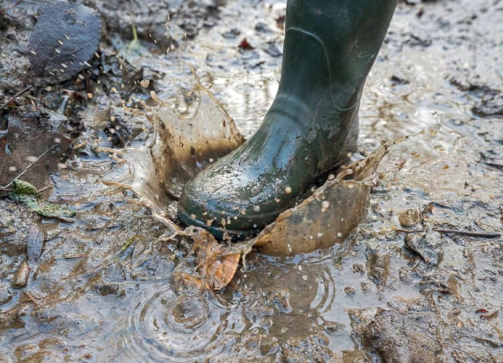 Gummistiefel tritt in schlammige Pfütze, Wasser und Matsch spritzen dynamisch nach oben.