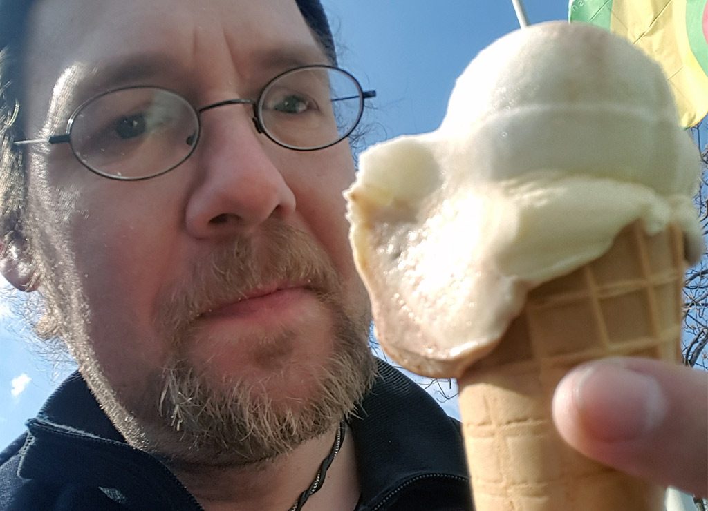 Mann mit Brille hält bei blauem Himmel eine Eiswaffel mit zwei hellen Kugeln in die Kamera.