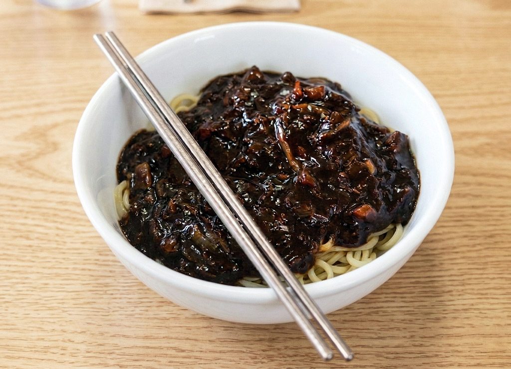 Schüssel mit Nudeln in schwarzer Bohnensauce, Essstäbchen liegen quer obenauf.