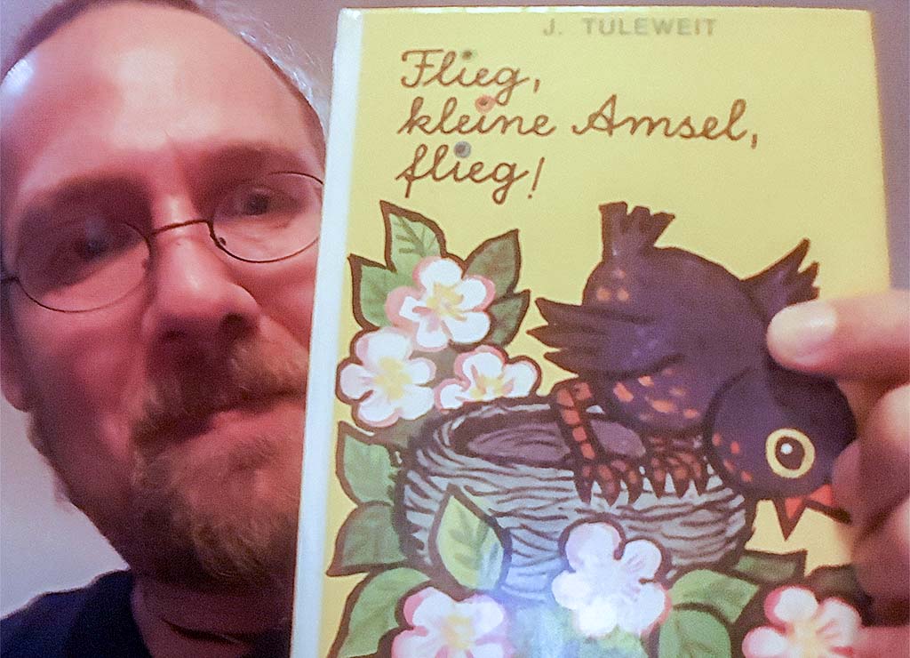 Internationaler Kinderbuchtag – International Children’s Book Day. Kuriose Feiertage - 2. April © 2024 Sven Giese – Bild 2 Mann mit Brille hält Kinderbuch „Flieg, kleine Amsel, flieg!“ mit gezeichneter Amsel ins Bild.