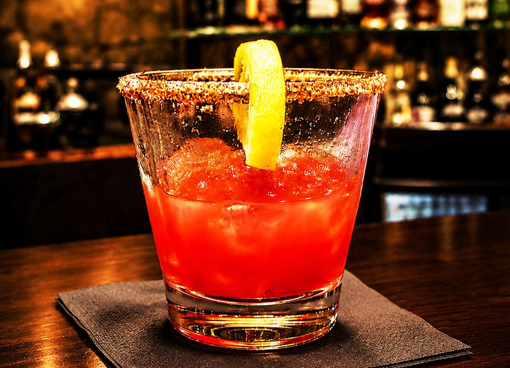 Roter Cocktail mit Zitronenscheibe und Gewürzrand auf Serviette vor Barhintergrund.