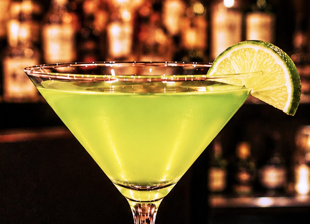 Grüner Cocktail im Martiniglas mit Limettenscheibe vor unscharfem Barhintergrund.