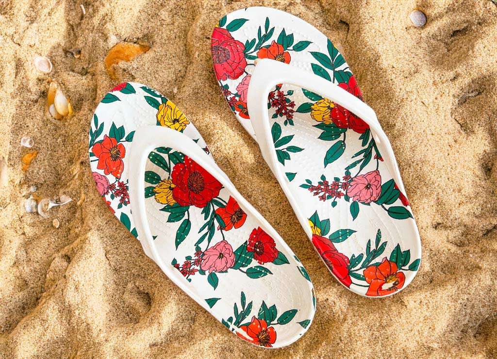 National Flip Flop Day – Tag der Flip-Flops in den USA. Kuriose Feiertage – dritter Freitag im Juni © 2024 Sven Giese - Bild 1 Weiß-bunte Flip-Flops mit Blumenmuster liegen auf Sand mit Muscheln.