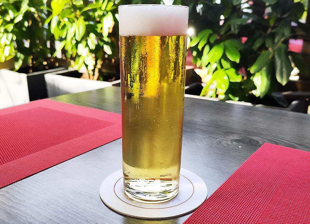 National Beer Lover’s Day – Tag der Bierliebhaber in den USA. Kuriose Feiertage - 7. September © 2024 Sven Giese - Bild 1 Ein Glas helles Bier mit Schaumkrone steht auf einem Bierdeckel auf einem Tisch, der mit roten Platzdeckchen gedeckt ist. Im Hintergrund sind grüne Pflanzen zu sehen.