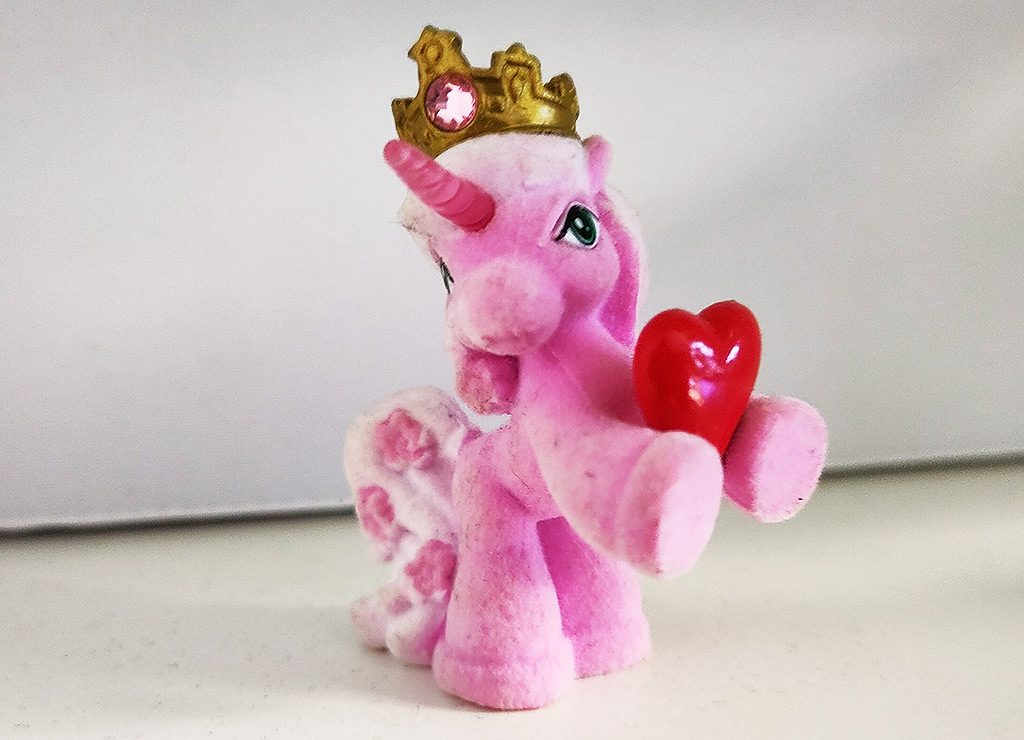 Ein rosafarbenes Spielzeug-Einhorn mit einer Krone, das aufrecht steht und ein rotes Herz zwischen den Vorderhufen hält.