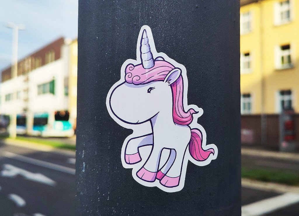 Ein Aufkleber eines gezeichneten Einhorns mit rosa Mähne und Horn auf einem grauen Laternenpfahl in einer städtischen Umgebung.