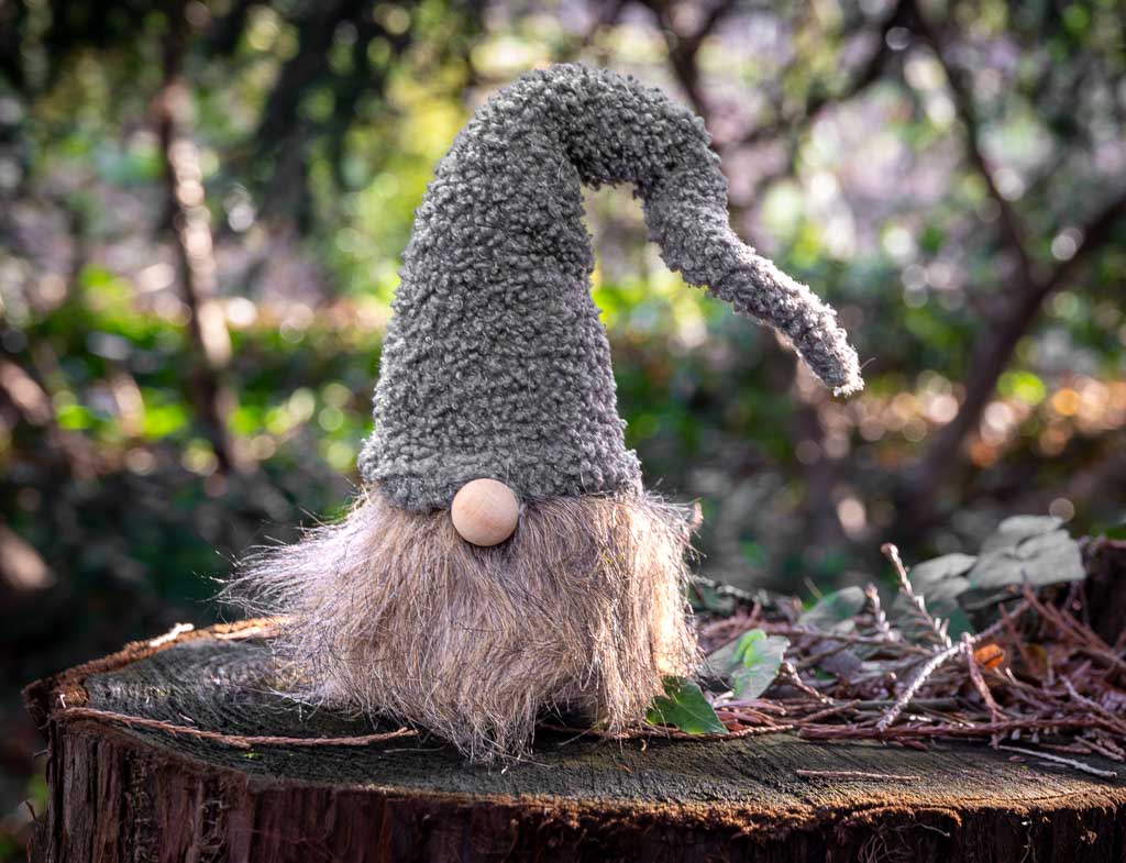 Waldmännchentag oder Schlägelstag in Deutschland. Kuriose Feiertage - 2. Januar © 2024 Sven Giese – Bild 2 Ein gestricktes Waldmännchen mit grauer Mütze und Holzknopfnase sitzt auf einem Baumstumpf im Wald.