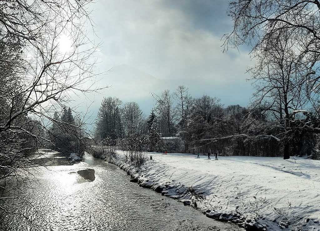 Verschneite Flussufer mit kahlen Bäumen und Spaziergängern im Hintergrund unter bewölktem Himmel.