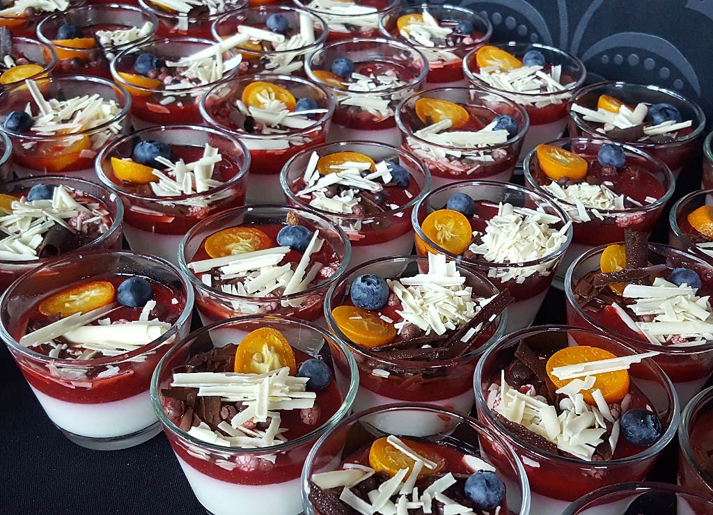 Eine Vielzahl von Dessertgläsern, gefüllt mit einer Schicht aus weißem Pudding und roter Fruchtsoße, garniert mit weißen Schokoladenspänen, Blaubeeren und Physalis-Hälften.