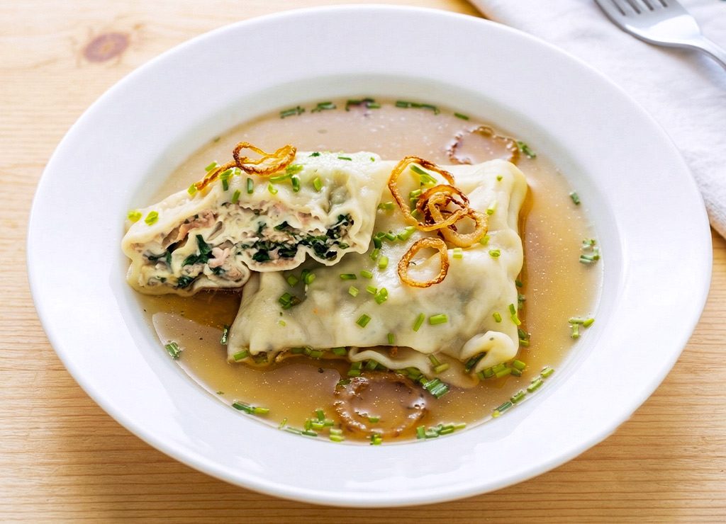 Weltmaultaschentag. Kuriose Feiertage – Gründonnerstag © 2026 Sven Giese - Bild 2 Maultaschen in Brühe auf weißem Teller, Schnittlauchstreu und Röstzwiebeln, Tisch im Hintergrund.