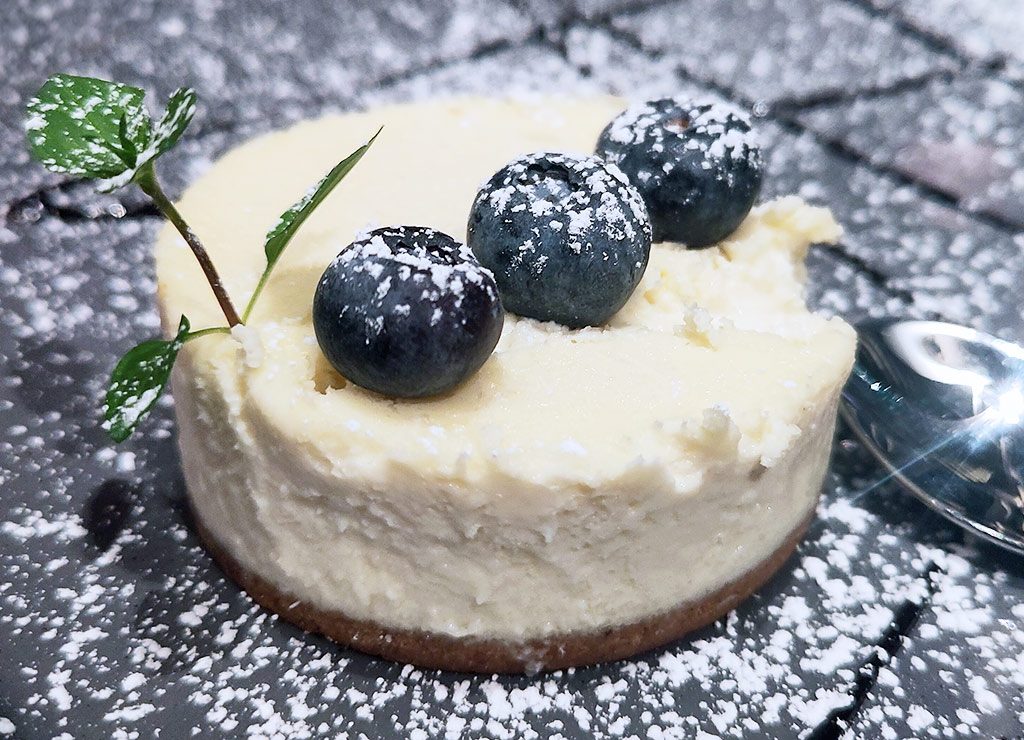 Blueberry Cheesecake Day – Tag des Blaubeerkäsekuchens in den USA. Kuriose Feiertage - 26. Mai © 2026 Sven Giese – Bild 1 Blaubeer-Käsekuchen mit Puderzucker und Minze auf dunklem Teller, Löffel daneben.