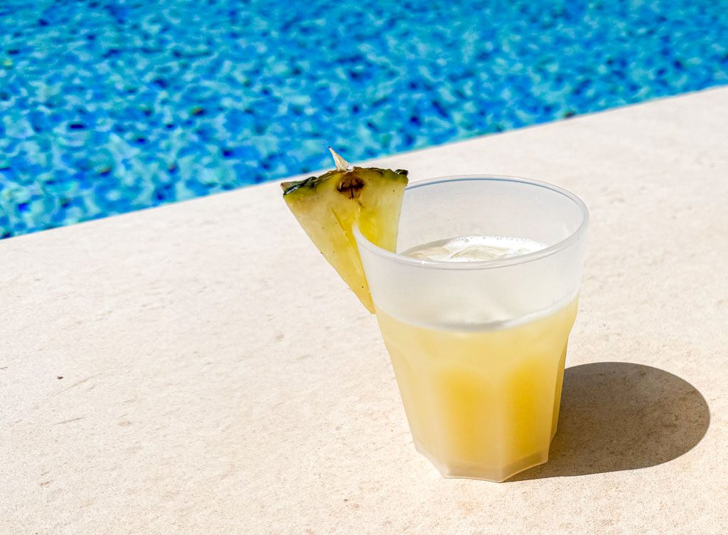 Gelber Ananas-Drink in einem Glas am Rand eines Pools mit blauem Wasser.