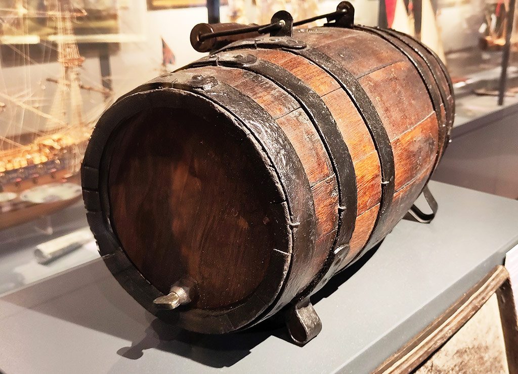 Ein antikes Rumfass aus Holz mit Metallringen und einem Zapfhahn, ausgestellt in einem Museum. Das Fass, das auf alten Schiffen genutzt wurde, ist auf einem Ständer platziert und scheint aus dunklem Holz gefertigt zu sein, mit deutlichen Gebrauchsspuren, die auf ein hohes Alter hinweisen. Im Hintergrund sind maritime Exponate und Modellschiffe zu sehen.