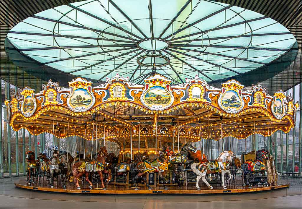 Jane's Carousel in Brooklyn, New York. Tag des Karussells in den USA © 2025 Sven Giese – Bild 1 Historisches Pferdekarussell mit bunter Beleuchtung hinter Glasfassade in moderner Halle.