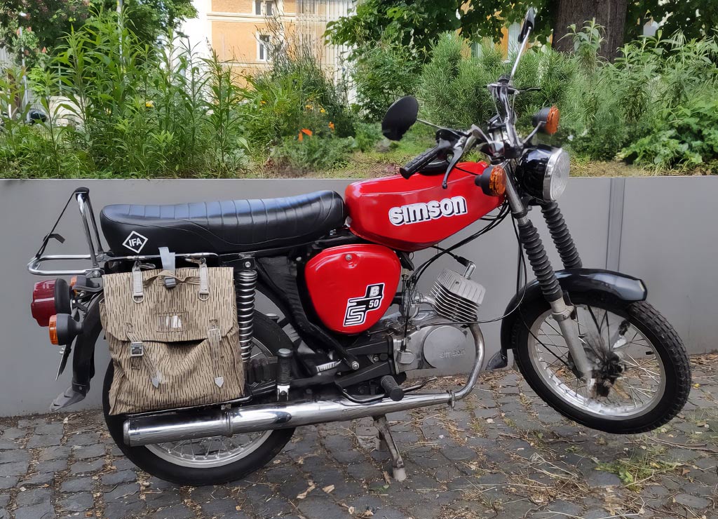 Simson-Tag in Deutschland. Kuriose Feiertage - 19. Juli © 2023 Sven Giese – Bild 5 Rote Simson S50 mit Packtasche parkt vor moderner grauer Mauer.