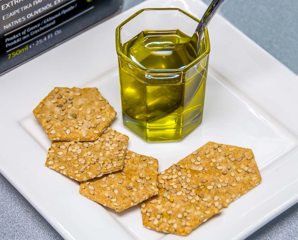 Ein Glas mit Olivenöl, umgeben von Sesam-Crackern auf einem weißen Teller.