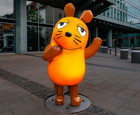 Große orange Maus-Figur winkt mit Pfote auf städtischem Platz vor Glasfassade und Pfeilern.