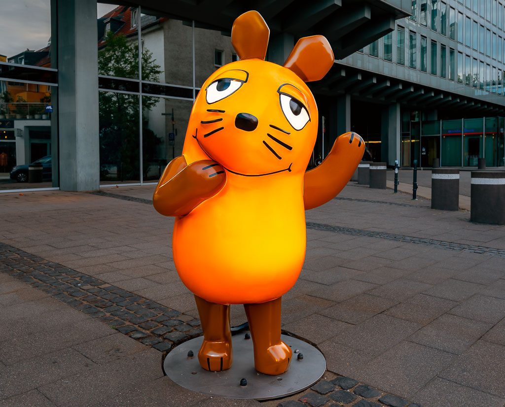 Große orange Maus-Figur winkt mit Pfote auf städtischem Platz vor Glasfassade und Pfeilern.