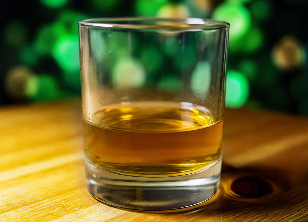 Glas mit irischem Whiskey auf Holztisch, warmes Licht, grüner Bokeh-Hintergrund im Abendambiente.