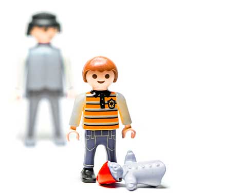 Playmobil-Kind mit gestreiftem Pullover und Puppe, eine erwachsene Figur im Hintergrund.