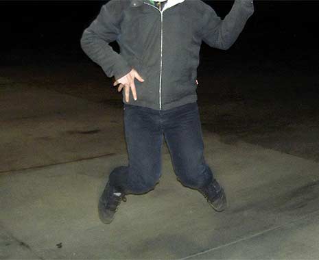 Person in dunkler Kleidung springt nachts auf Betonfläche in die Höhe, Gesicht nicht sichtbar.