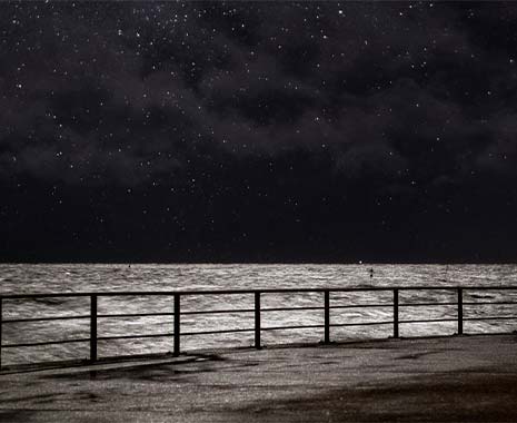 Ein nächtlicher Blick auf das Meer, das im silbrigen Mondlicht glitzert. Im Vordergrund ist ein Geländer zu sehen, dahinter die unruhige Wasseroberfläche und der sternenklare Himmel im Hintergrund.