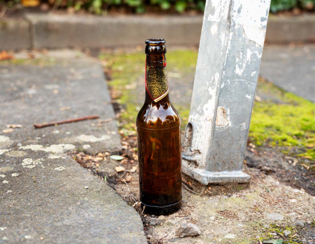 Leere Bierflasche neben Metallpfosten, auf einem leicht vermoosten und grauen Gehweg.