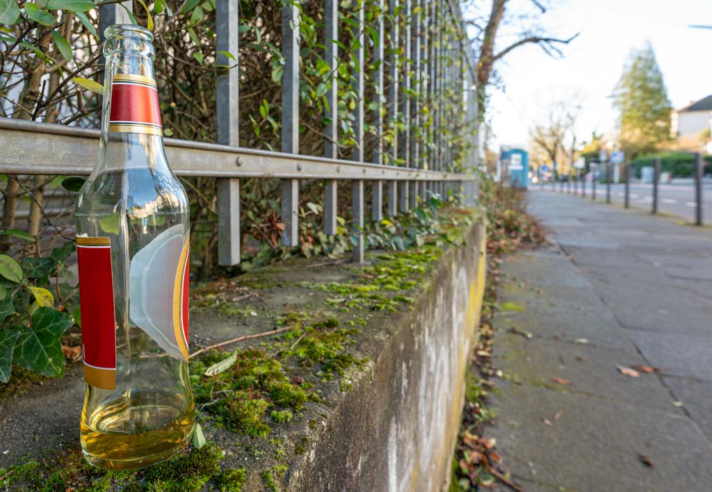 Durchsichtige Bierflasche mit rotem Etikett, auf vermooster Mauer an einem Metallzaun.