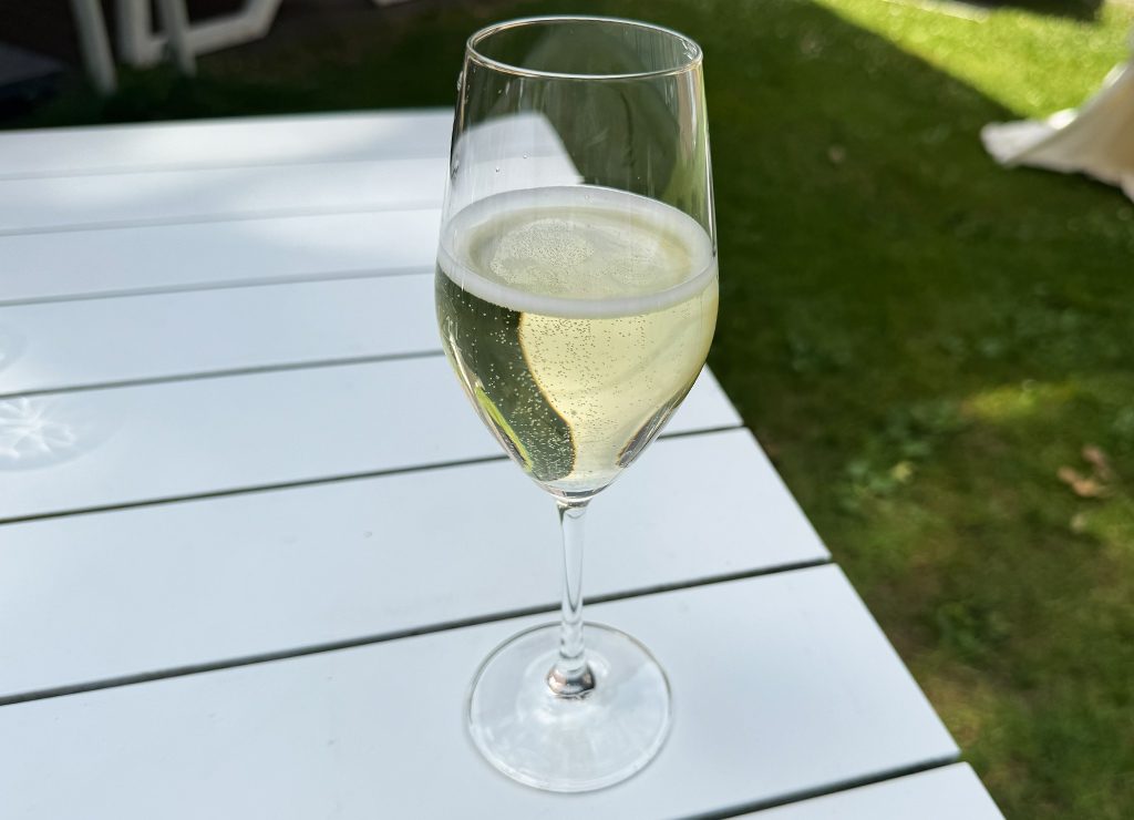 Gefülltes Sektglas steht auf weißem Gartentisch, im Hintergrund liegt eine sonnige Rasenfläche.