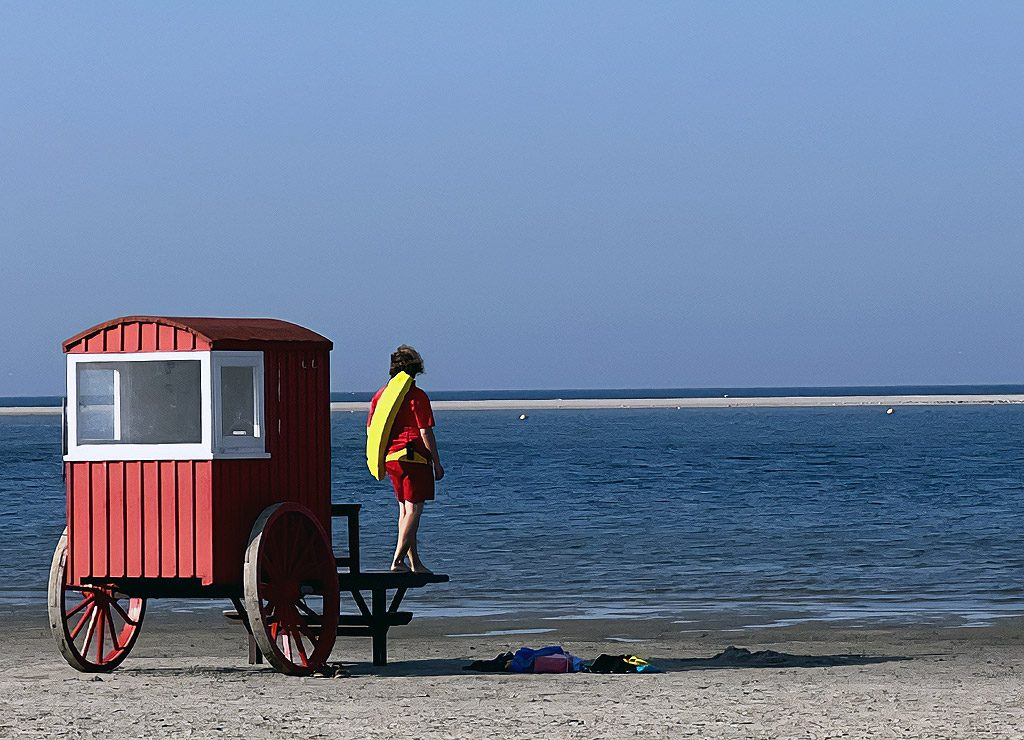 International Lifeguard Appreciation Day – internationaler Ehrentag der Rettungsschwimmer. Kuriose Feiertage – 31. Juli © 2026 Sven Giese Rettungsschwimmerin mit gelber Rettungsboje steht neben rotem Wachhäuschen und blickt aufs Meer.