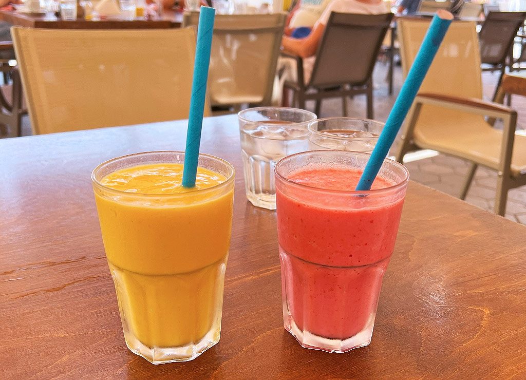 Ein roter und ein gelber Smoothie in Gläsern mit Strohhalm stehen auf einem Holztisch in einem Café. 