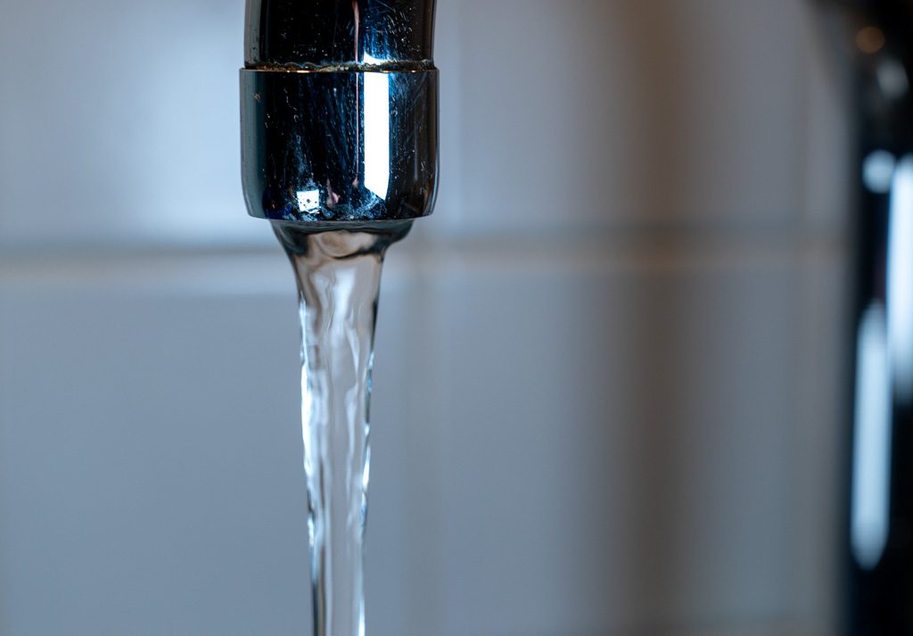 Fließendes Wasser aus einem glänzenden, metallischen Wasserhahn vor hellen Fliesen.