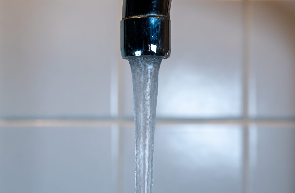 Fließendes Wasser aus einem metallischen Wasserhahn vor neutralem Hintergrund mit Fliesen.