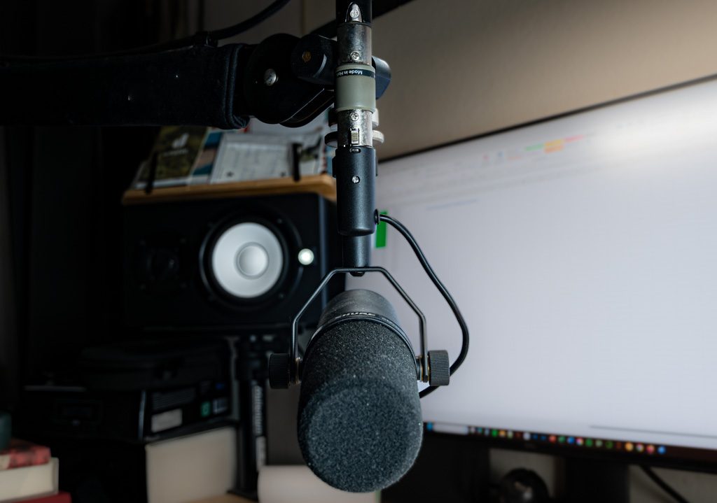 Ein Studiomikrofon in einem Raum mit Lautsprecher, Monitor und Audioequipment im Hintergrund.