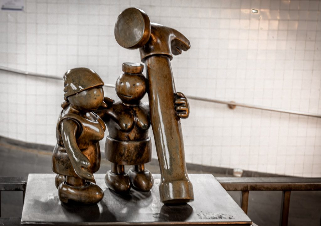Bronzestatue zweier Figuren mit einem übergroßen Hammer, ausgestellt in einem U-Bahn-Bereich.