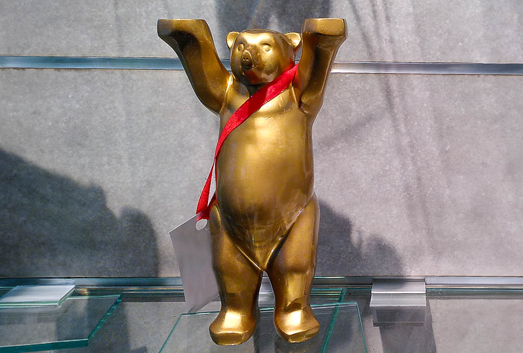 Tag des Berliner Bären. Kuriose Feiertage - 22. März © 2024 Sven Giese. Goldene Bärenfigur mit erhobenen Armen und rotem Band um den Hals vor grauem Hintergrund.
