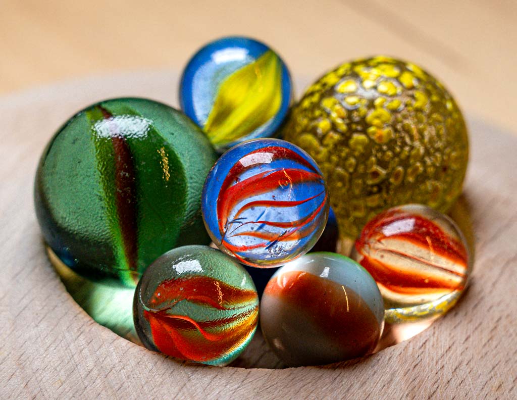 World Marbles Day – Welt-Murmel-Tag. Kuriose Feiertage – immer am Karfreitag © 2024 Sven Giese – Bild 1 Mehrere bunte Glasmurmeln liegen eng zusammen in einer runden Vertiefung aus Holz.