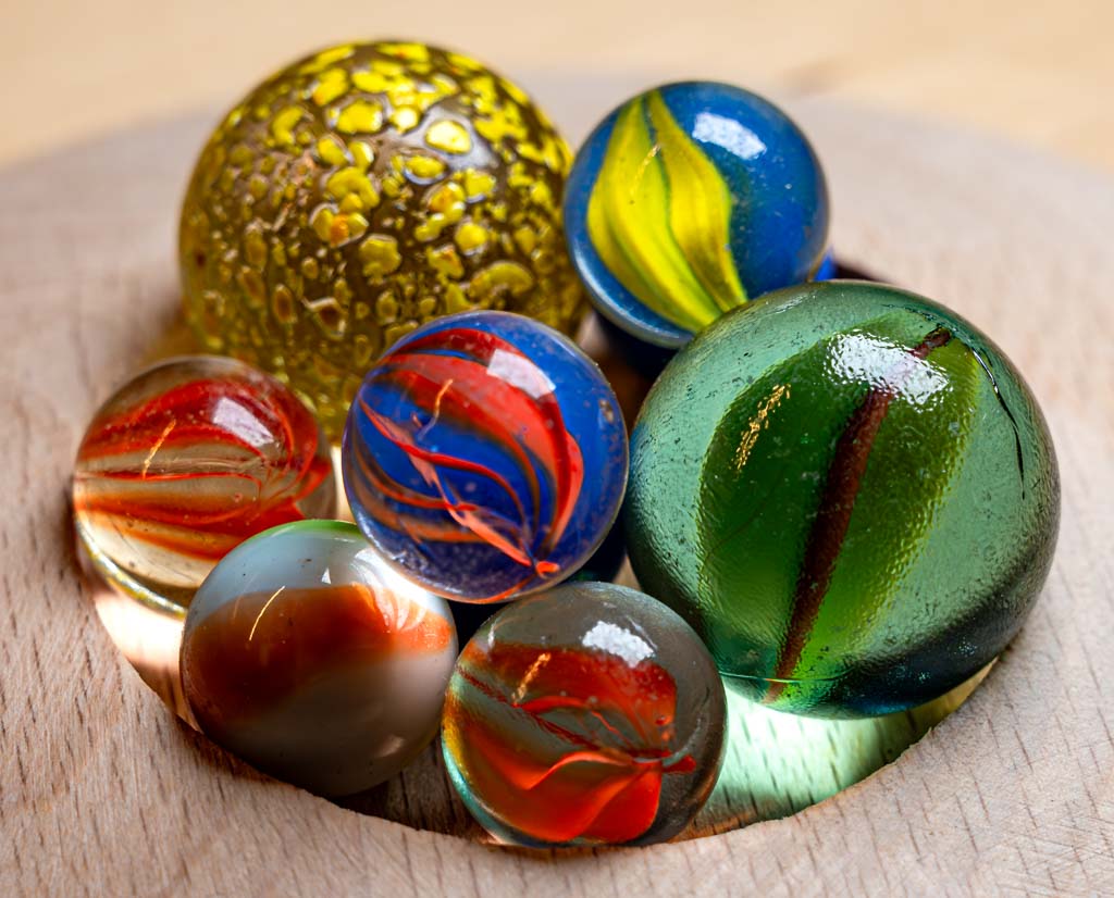 World Marbles Day – Welt-Murmel-Tag. Kuriose Feiertage – immer am Karfreitag © 2024 Sven Giese – Bild 3 Mehrfarbige Glasmurmeln mit Mustern in einer Vertiefung auf hellem Holz arrangiert.