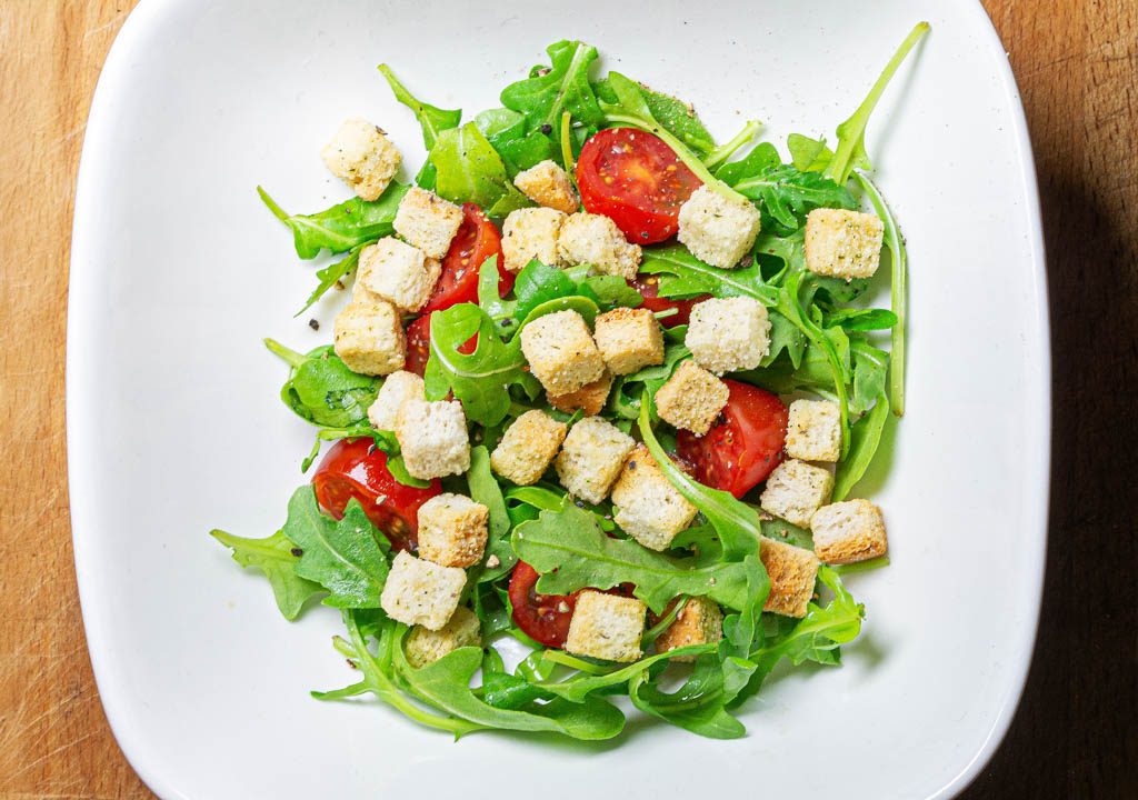 Frischer Rucolasalat mit Tomaten und knusprigen Croutons in einem weißen Teller serviert.
