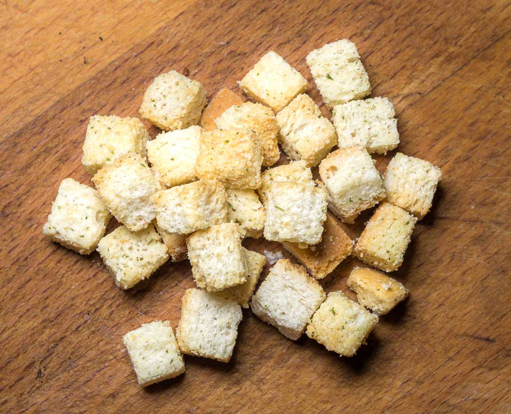 Mehrere geröstete Croutons mit Kräutern liegen locker verteilt auf einer Holzfläche.