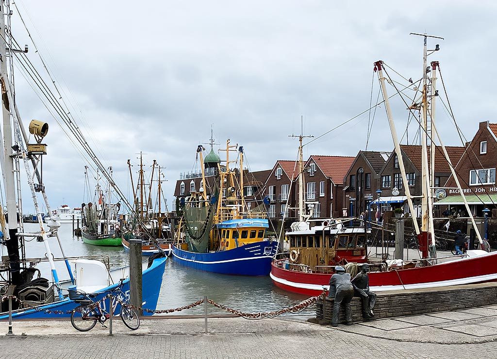 Fischerboote im Hafen mit roten Dächern im Hintergrund und zwei Bronzefiguren im Vordergrund.