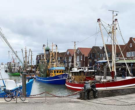 Fischerboote im Hafen mit roten Dächern im Hintergrund und zwei Bronzefiguren im Vordergrund
