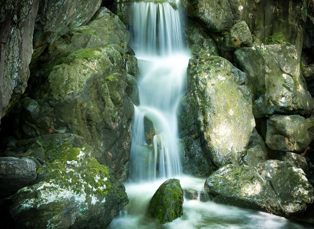 Kleiner Wasserfall, der zwischen moosbewachsenen Felsen in ein Becken herabstürzt.