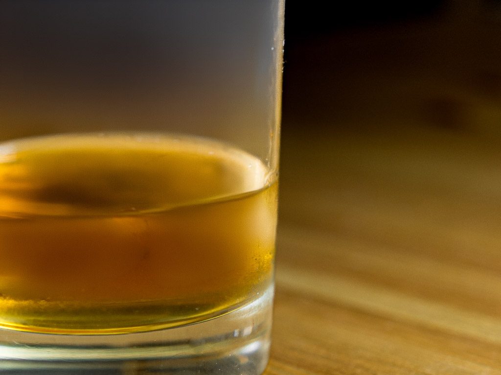 Halbvolles Glas mit bernsteinfarbenem Scotch, weich fokussiert auf Holztisch.